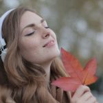 Wireless Audio - Young satisfied woman in headphones with fresh red leaf listening to music with pleasure while lounging in autumn park