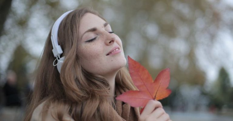 Wireless Audio - Young satisfied woman in headphones with fresh red leaf listening to music with pleasure while lounging in autumn park
