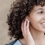 Bluetooth And Wi-Fi Audio - Cheerful ethnic female with black curly hair looking away with smile while listening to songs in wireless earphones on street against blurred background