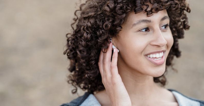 Bluetooth And Wi-Fi Audio - Cheerful ethnic female with black curly hair looking away with smile while listening to songs in wireless earphones on street against blurred background