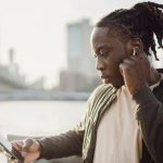 Wireless Audio Security - Thoughtful adult African American guy in wireless earphones listening to music while surfing internet on cellphone and standing leaned on hand near fence on bridge over city river