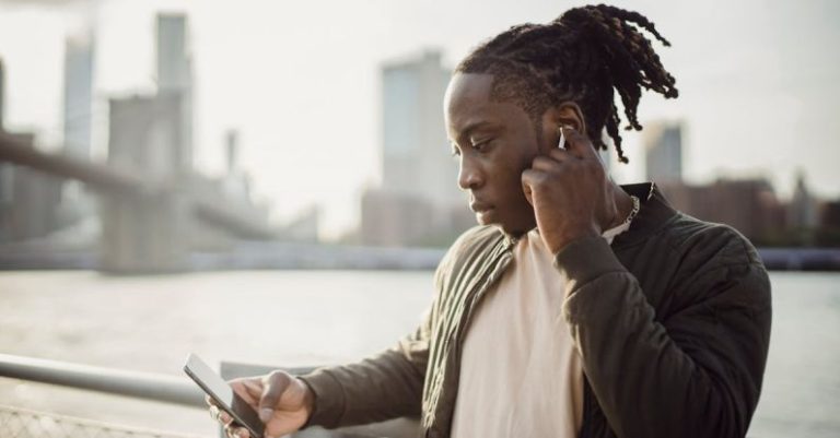 Wireless Audio Security - Thoughtful adult African American guy in wireless earphones listening to music while surfing internet on cellphone and standing leaned on hand near fence on bridge over city river