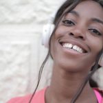 Wireless Audio - Positive young black female in bright t shirt wearing headphones and listening to music while looking at camera