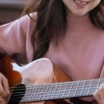 Room Acoustics - Smiling Asian female musician in casual wear playing acoustic guitar while sitting near piano during rehearsal at home in light room