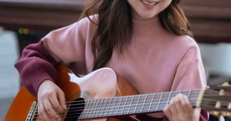 Room Acoustics - Smiling Asian female musician in casual wear playing acoustic guitar while sitting near piano during rehearsal at home in light room