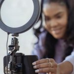 Podcast Platforms - Cheerful young African American female blogger in stylish sweater smiling while setting up camera of smartphone attached to tripod with ring light before recording vlog