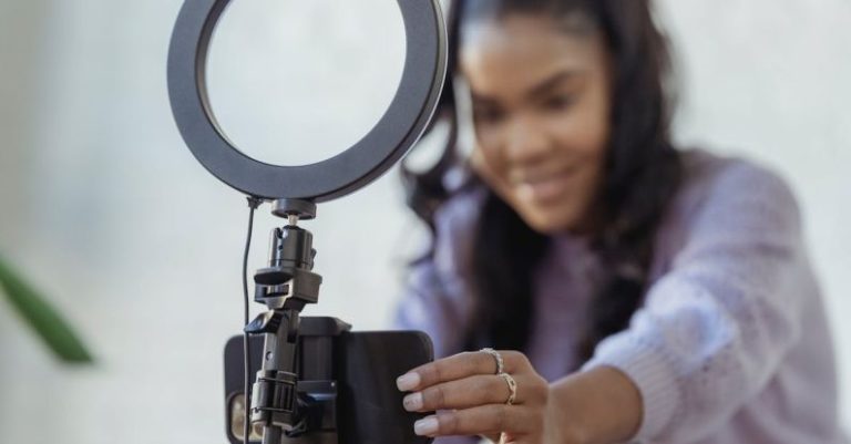 Podcast Platforms - Cheerful young African American female blogger in stylish sweater smiling while setting up camera of smartphone attached to tripod with ring light before recording vlog