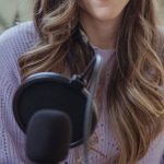 Podcast Content - Cheerful female radio host looking at camera while sitting in chair with opened notebook near professional microphone in light room
