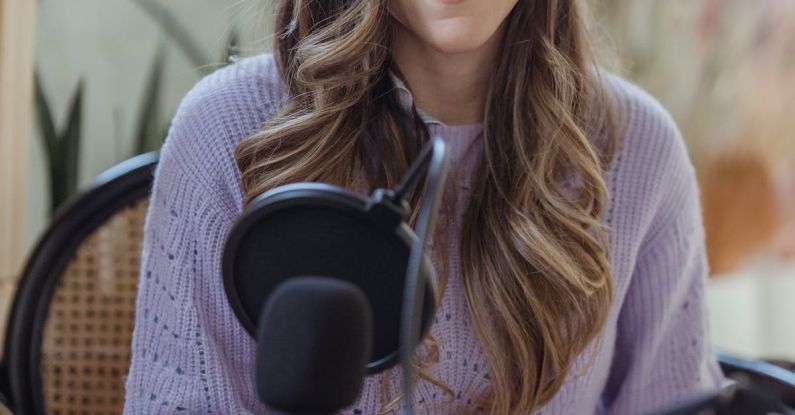 Podcast Content - Cheerful female radio host looking at camera while sitting in chair with opened notebook near professional microphone in light room