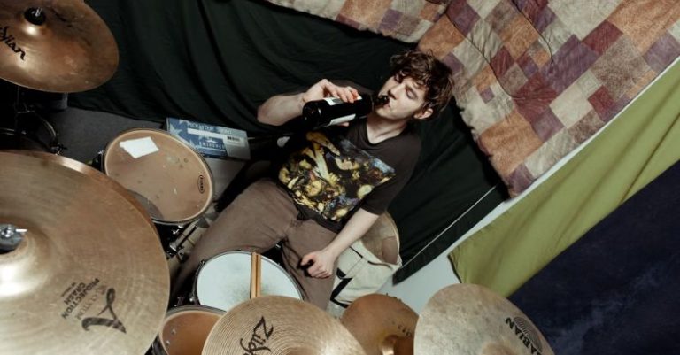 Soundproofing - From above of male musician in casual outfit sitting at drum set near wall with bottle of beer in studio